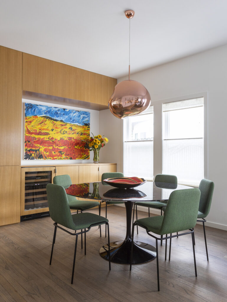 A modern dining room featuring a sleek black round dining table surrounded by six green chairs, as well as a metallic pendant light and a large colorful art piece.
